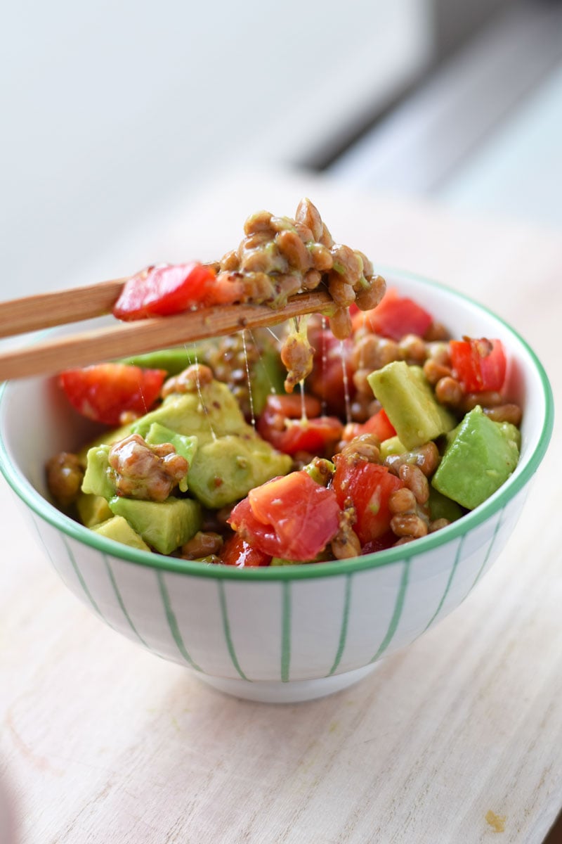 Natto with tomato and avocado, lifted from the bowl with sticky, gooey strings trailing down; dressed with olive oil-based salad dressing.