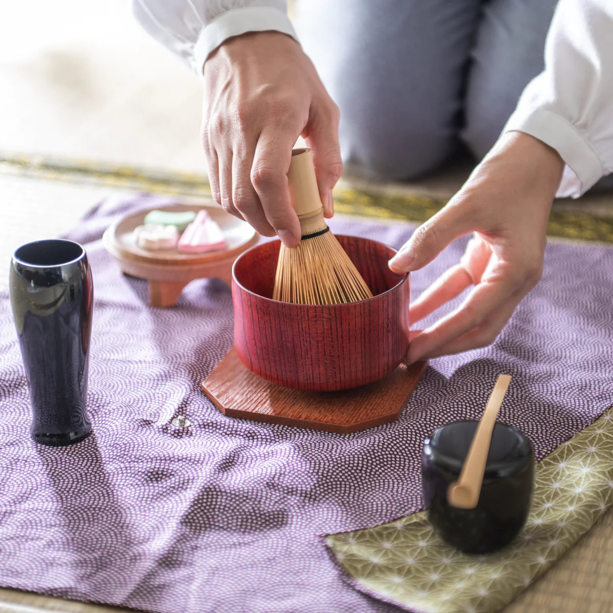 a person whisking matcha. She is kneeling on a tatami floor and holding the chawan tea bowl and bamboo whisk