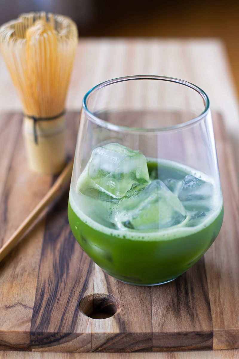matcha in glass with ice cubes with whisk in the background