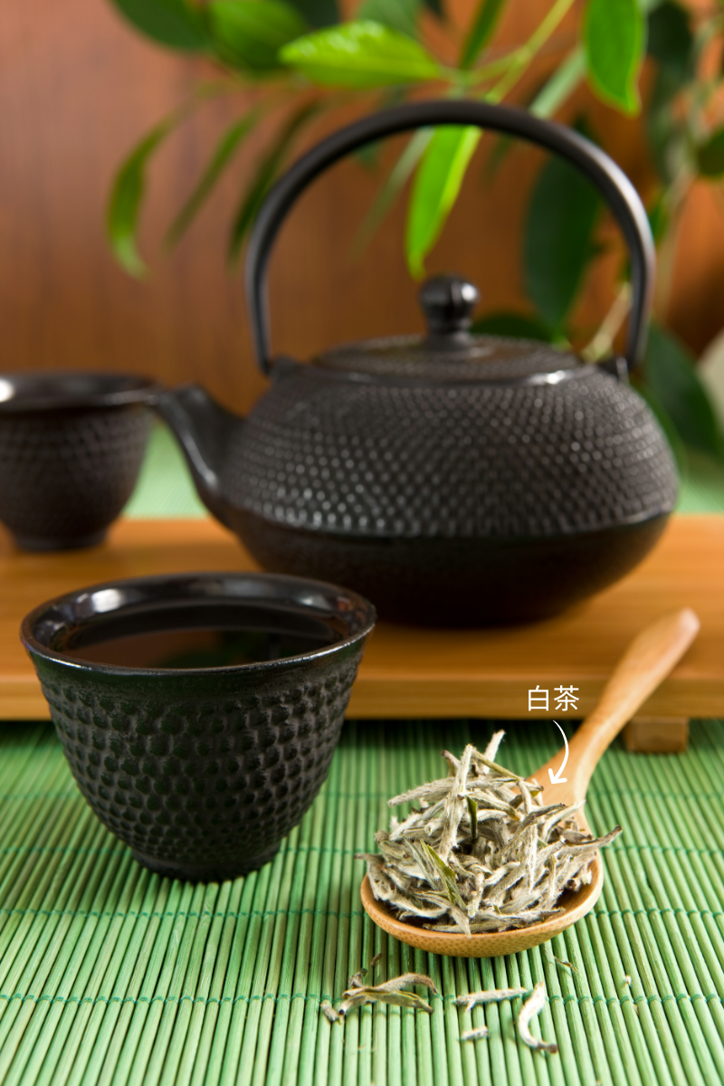 spoon of white tea leaves next to black tea cup and black iron tea pot in background