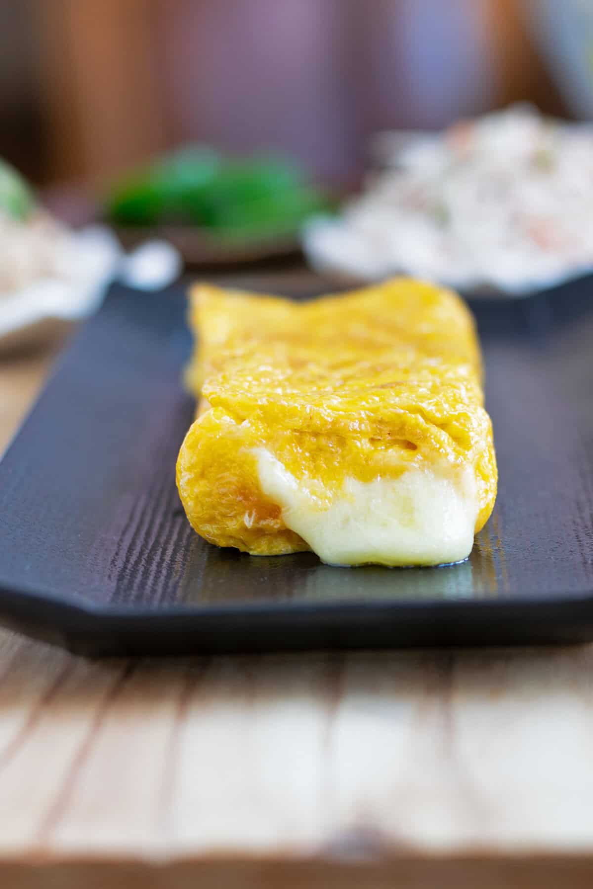 Tamagoyaki Japanese rolled omelet with warm mozzarella cheese oozing out the end onto a black serving plate.