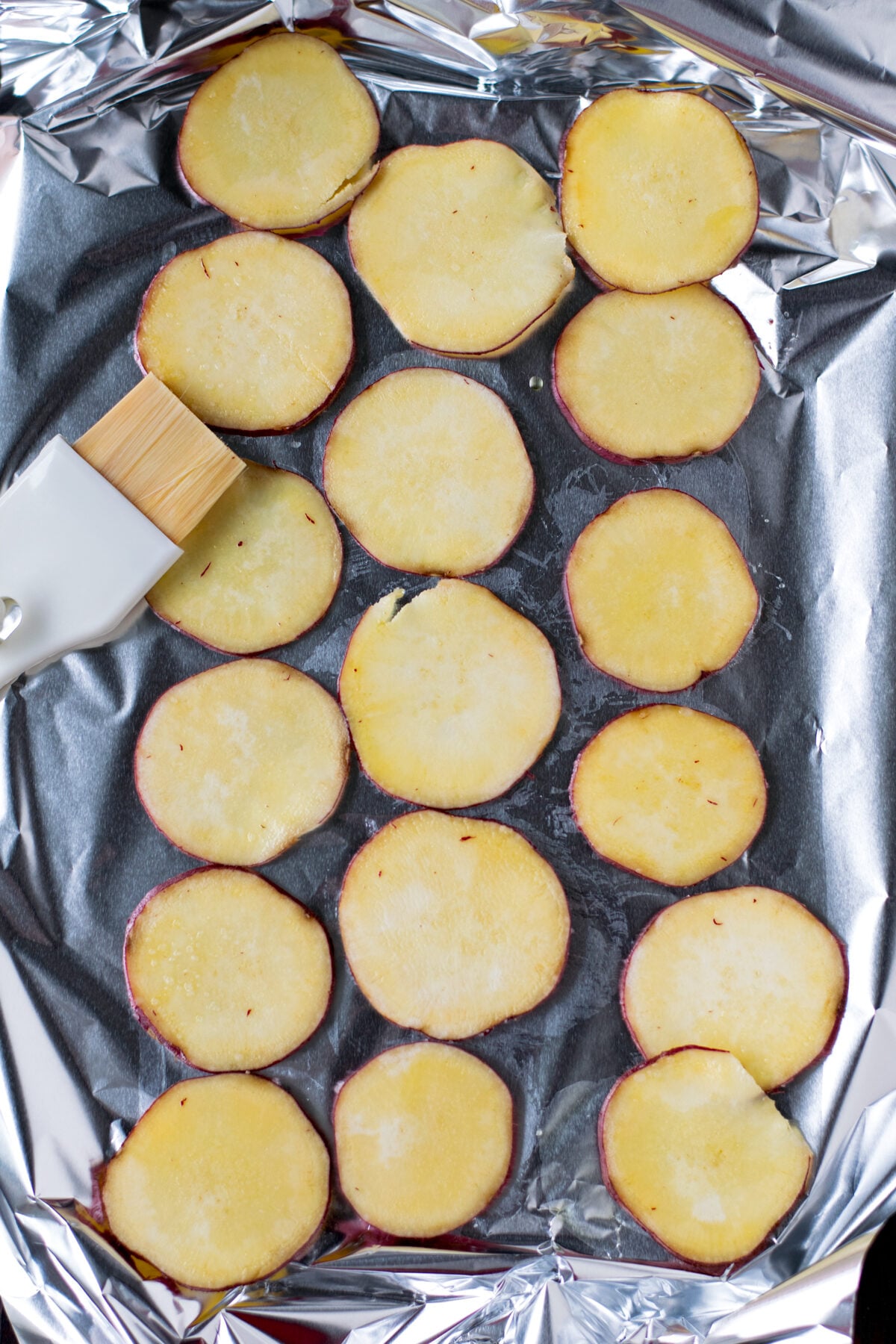 Slices of Japanese sweet potato on sheet of aluminum foil, coated with olive oil with brush, 