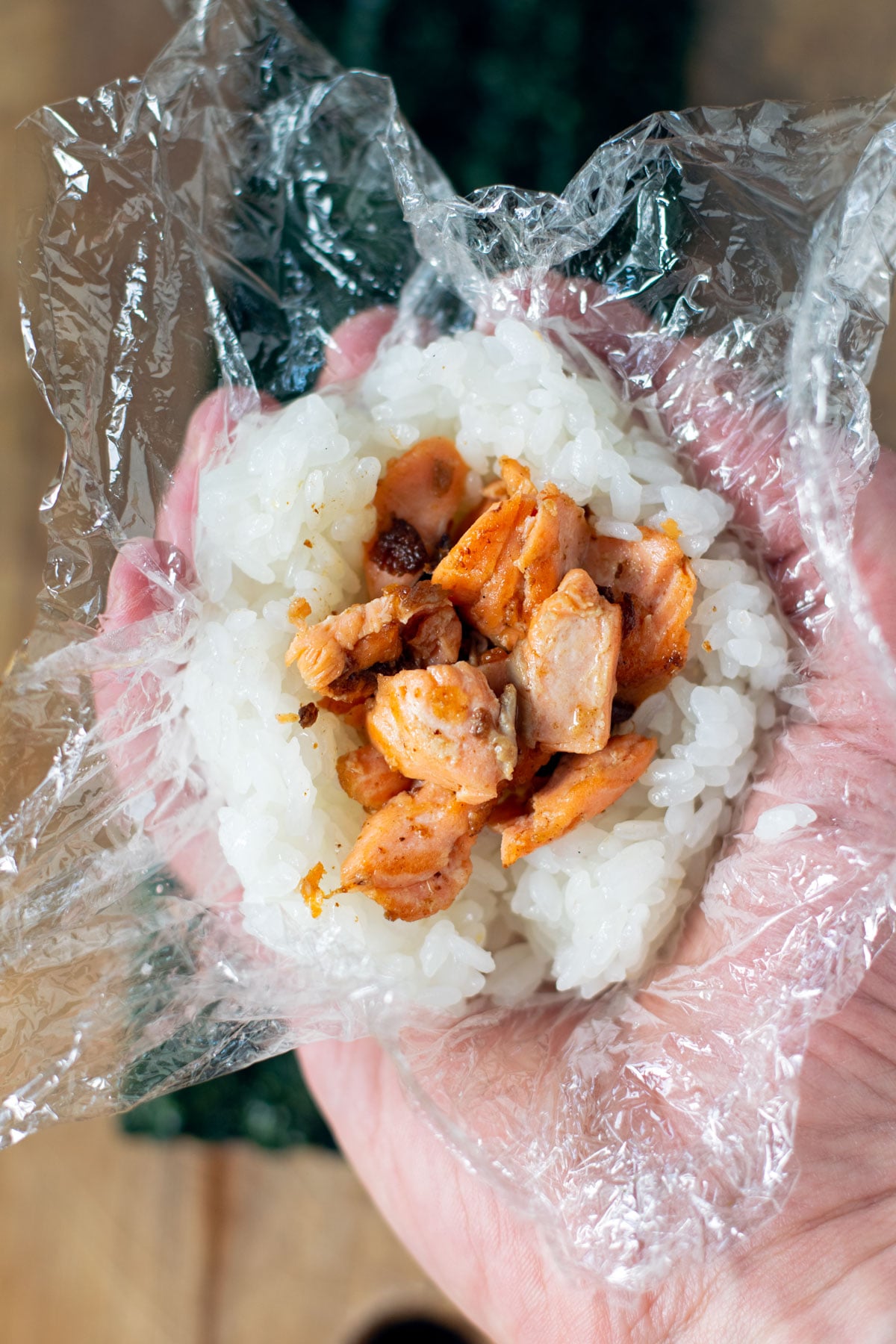 A hand holding a rice ball that is filled with grilled salmon and is wrapped with plastic wrap.