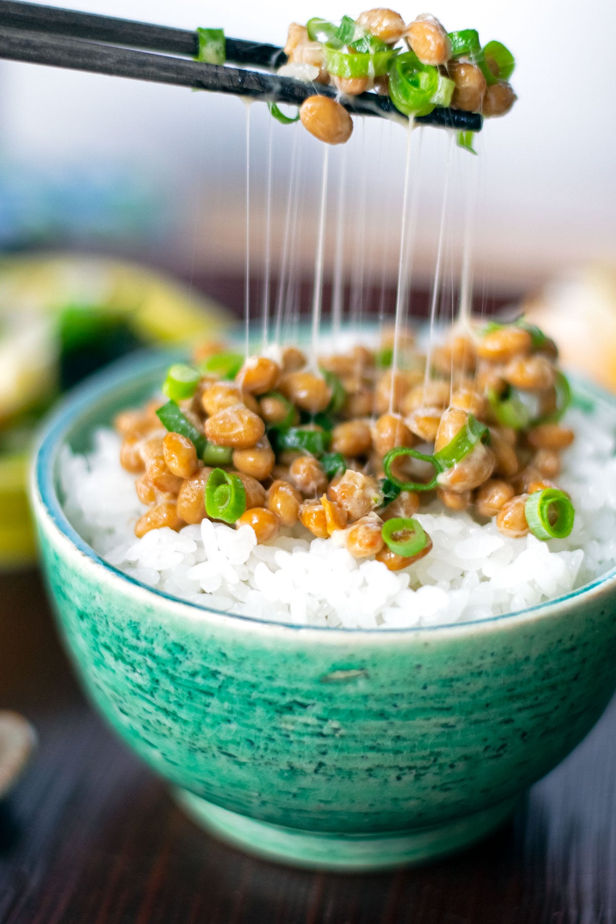 Chopsticks pulling up a bite of natto and sliced negi green onions with gooey strings stretching down to the bowl of white rice.
