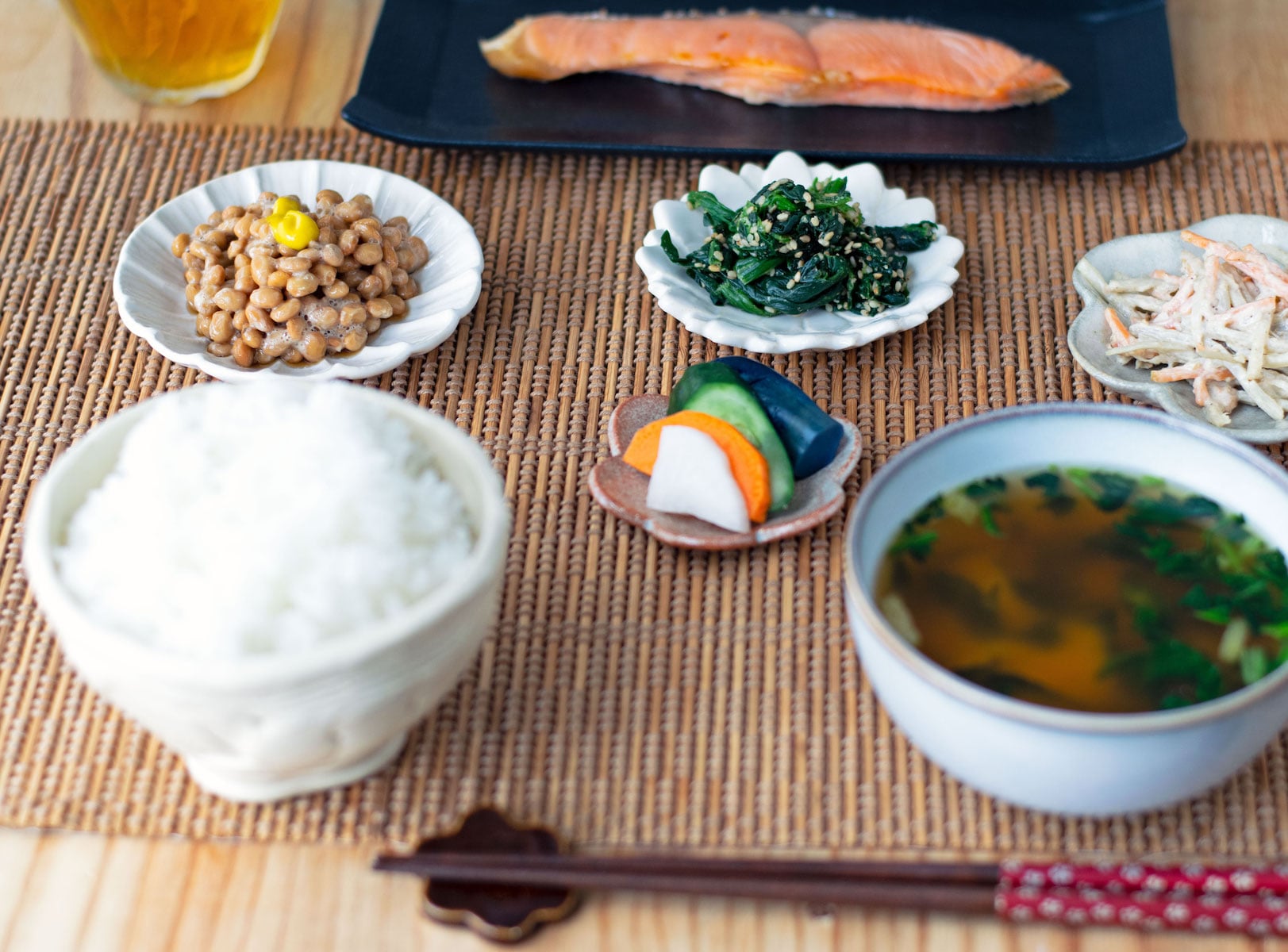 A full Japanese traditional breakfast with white rice, miso soup, pickles, spinach, burdock carrot salad, grilled salmon, and natto topped with karashi mustard.