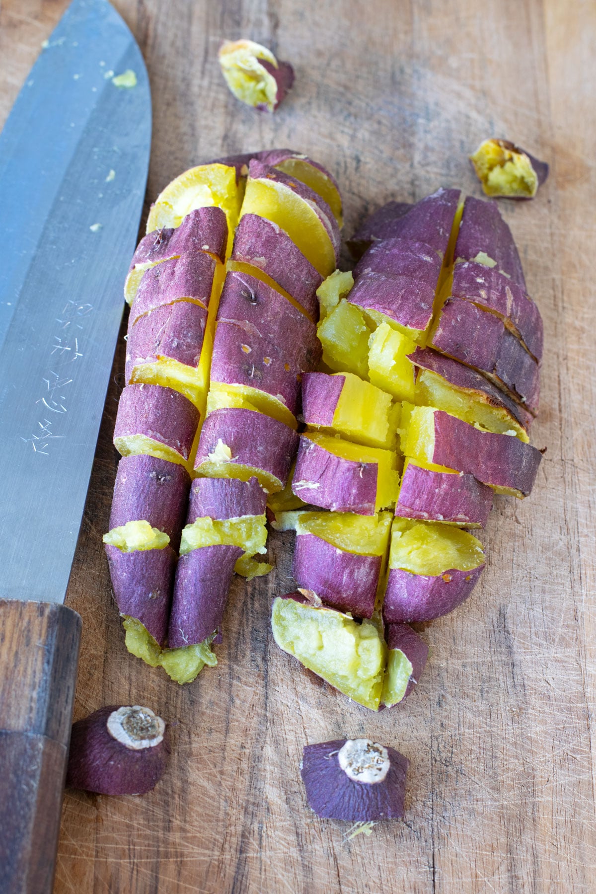 Two baked Japanese sweet potatoes sliced into small pieces on a wooden cutting board with a knife.