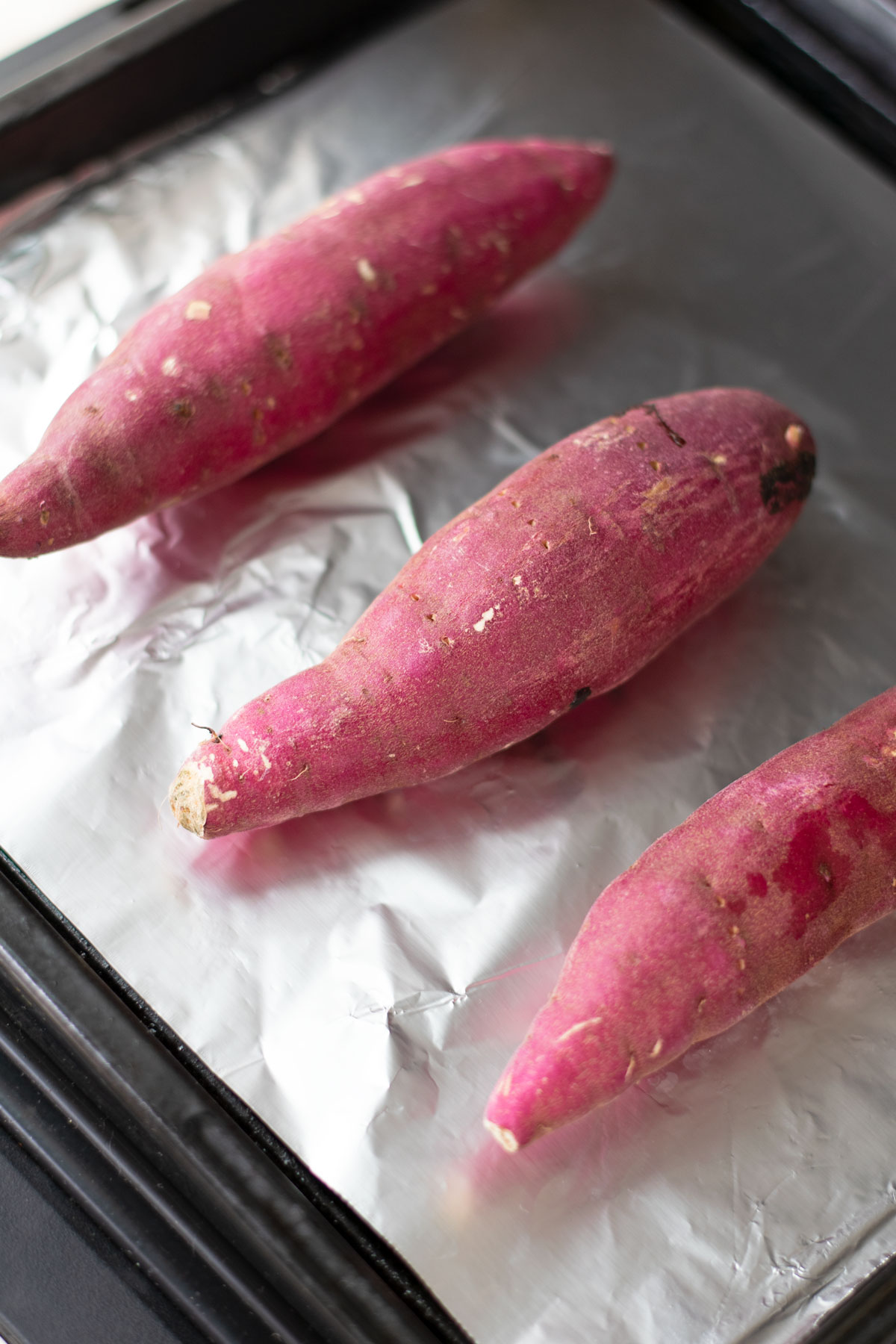 3 Japanese sweet potatoes on a baking sheet with foil going into the oven.