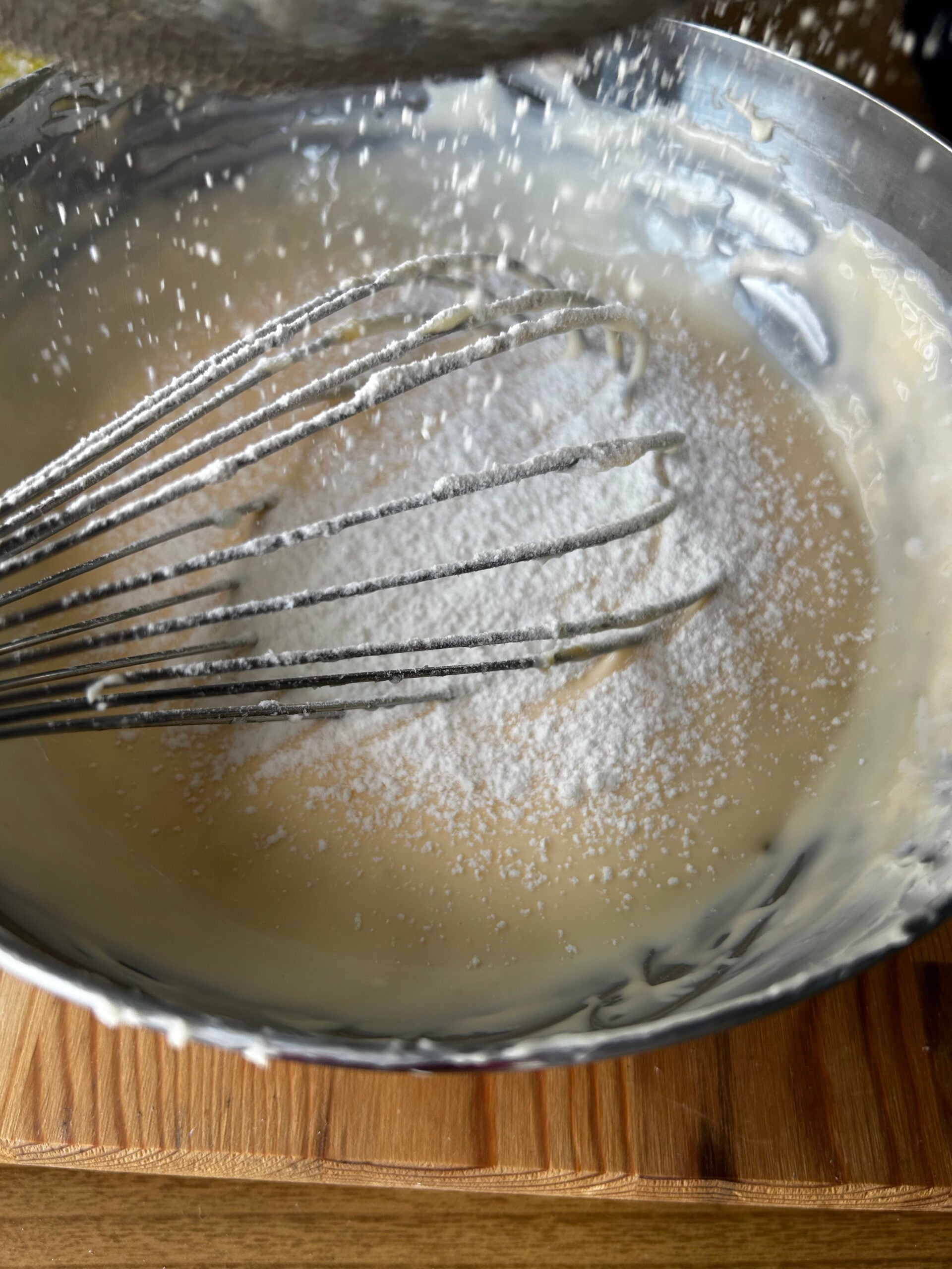 Sifting cake flour into cheesecake batter with a whisk inside the metel mixing bowl.