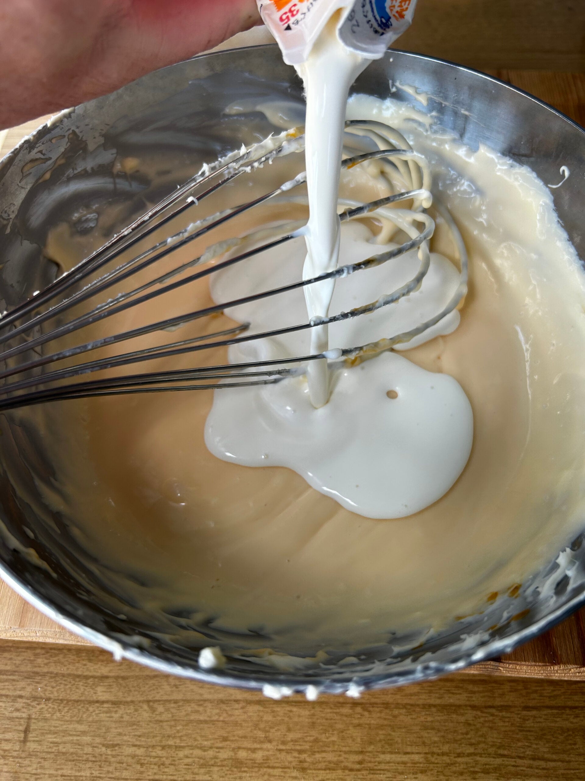 Pouring heavy cream into cheesecake batter in a metal mixing bowl.