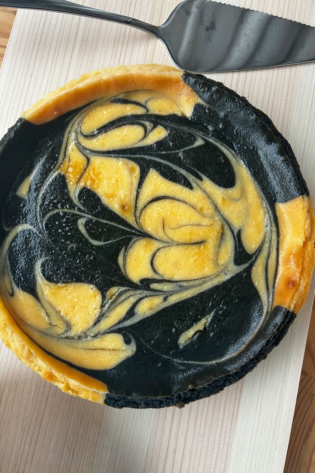 Whole marbled black sesame cheesecake on a cutting board with metal server.