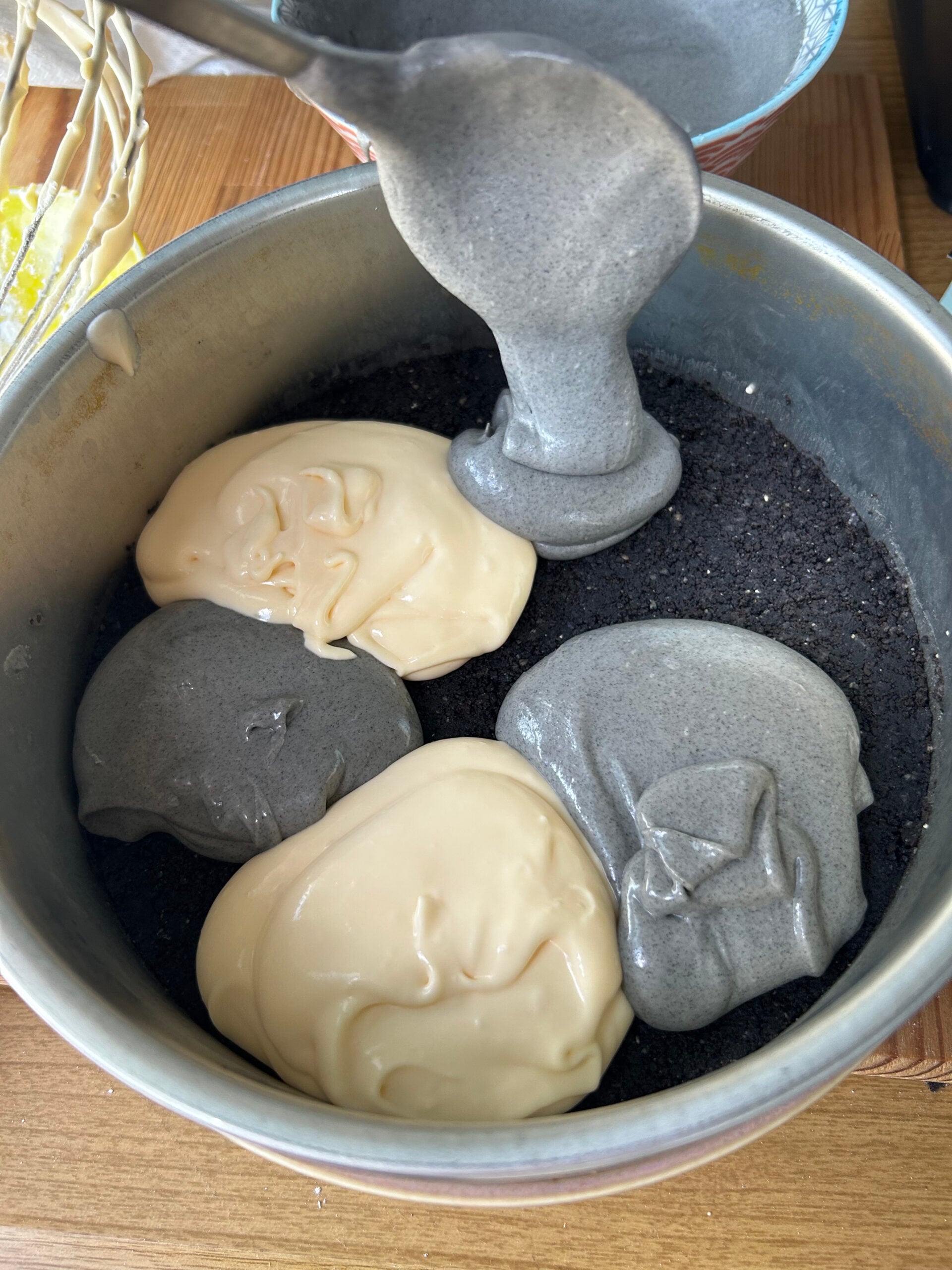 Spooning dollops or black sesame and plain cheesecake batter onto Oreo crust in a springform pan.